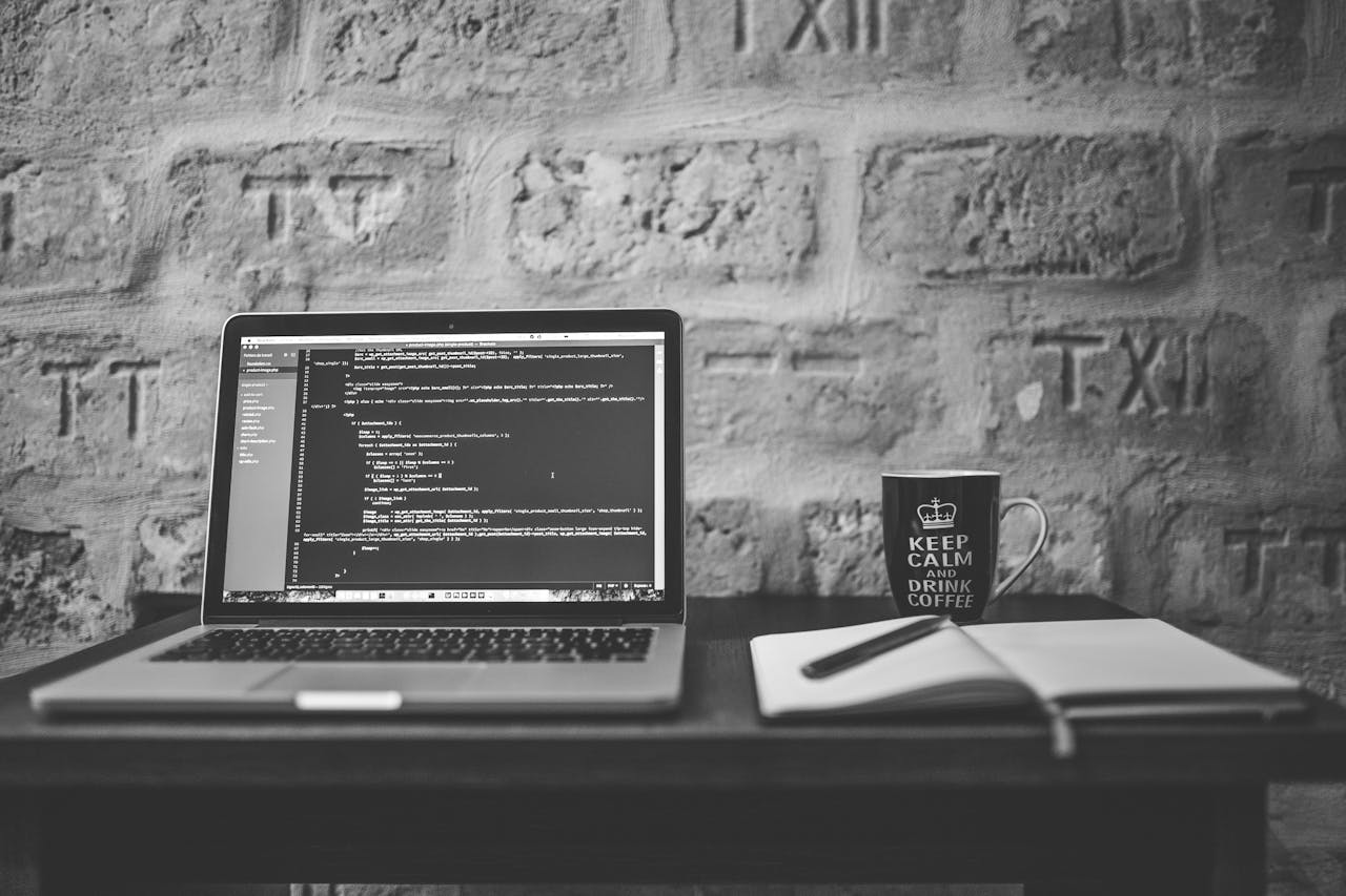 Black and white photo of a coding workspace with a laptop, mug, and notebook.